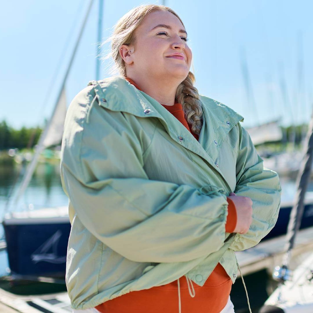 Frau mit geflochtenem Haar steht entspannt in einer Hafenumgebung. Sie trägt eine mintgrüne Jacke über einem orangefarbenen Oberteil. Im Hintergrund sind Boote und Masten zu sehen, die eine ruhige und maritime Atmosphäre schaffen.