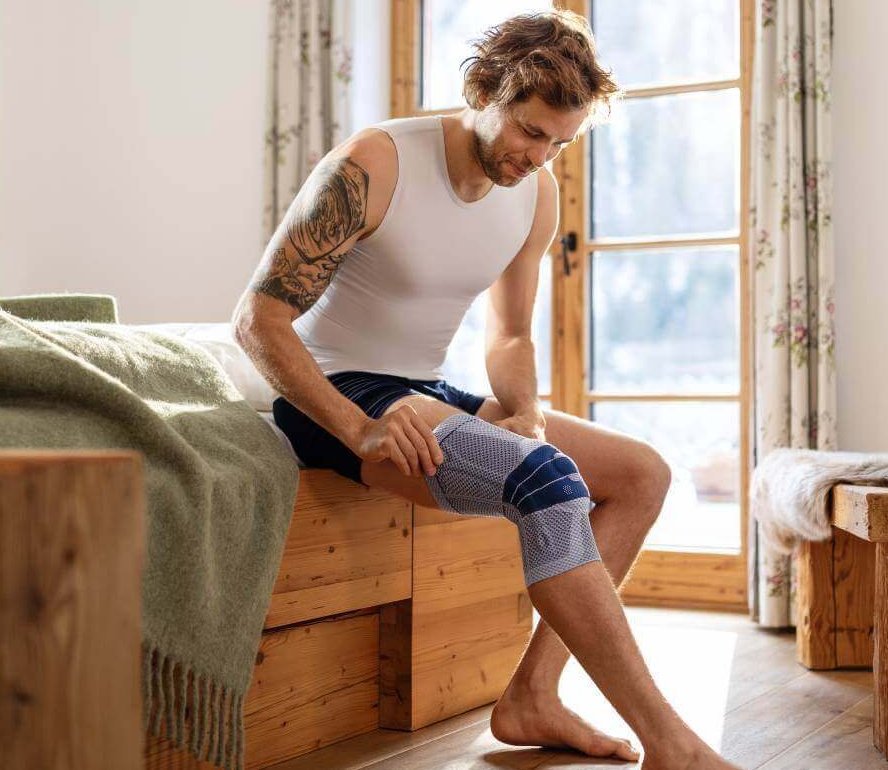 Ein junger Mann sitzt auf einem Bett um zieht eine Bandage an. Bei dieser handelt es sich um die GenuTrain P3 von Bauerfeind, die zur Behandlung von Knieschmerzen eingesetzt wird.