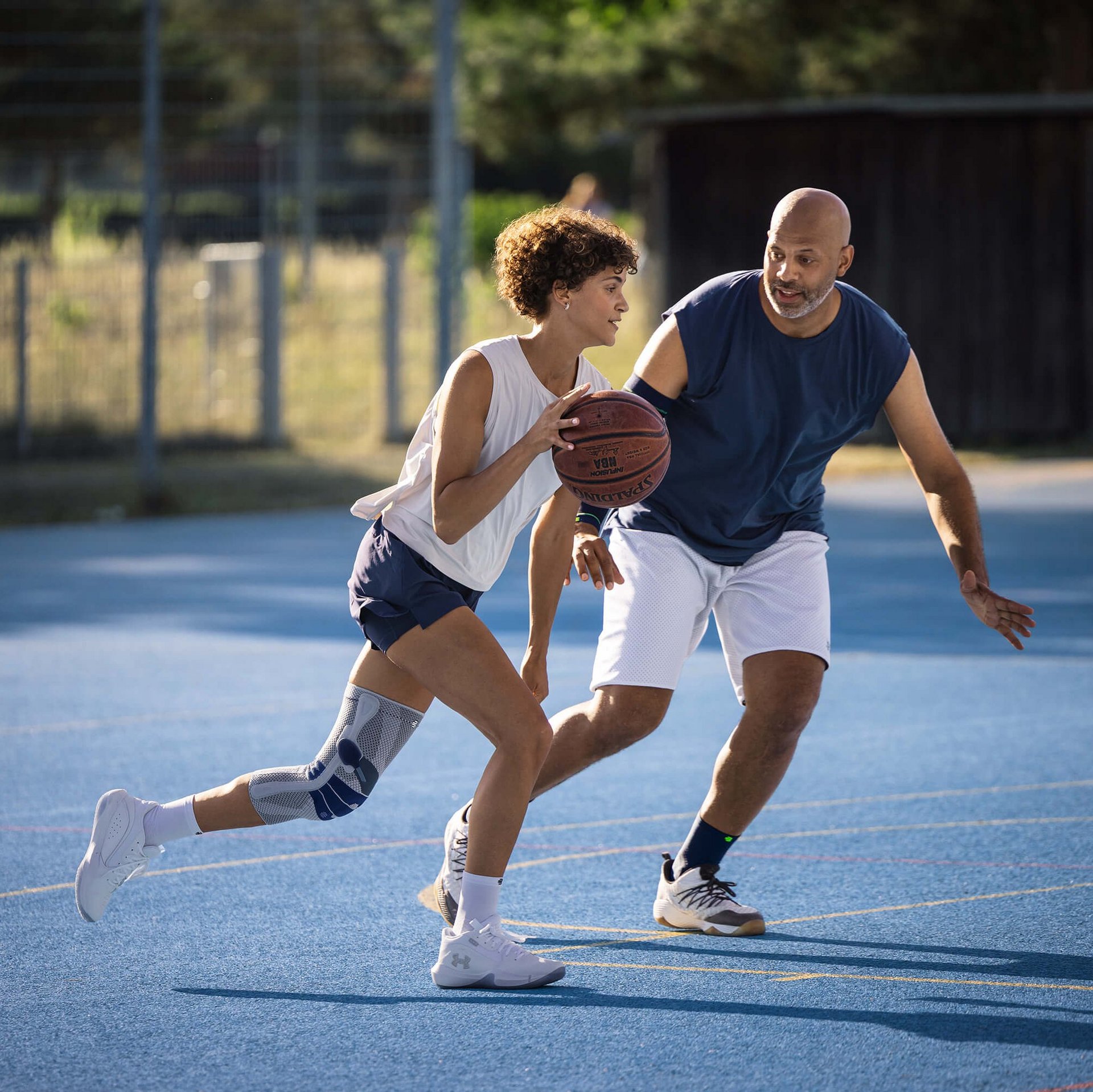 Zwei Personen spielen Basketball auf einem Aussenplatz mit blauem Bodenbelag. Die spielende Person im Vordergrund trägt eine Kniebandage am linken Bein und dribbelt den Ball, während die andere Person in Verteidigungsposition vor ihr steht. Beide sind sportlich gekleidet und konzentriert im Spielgeschehen.