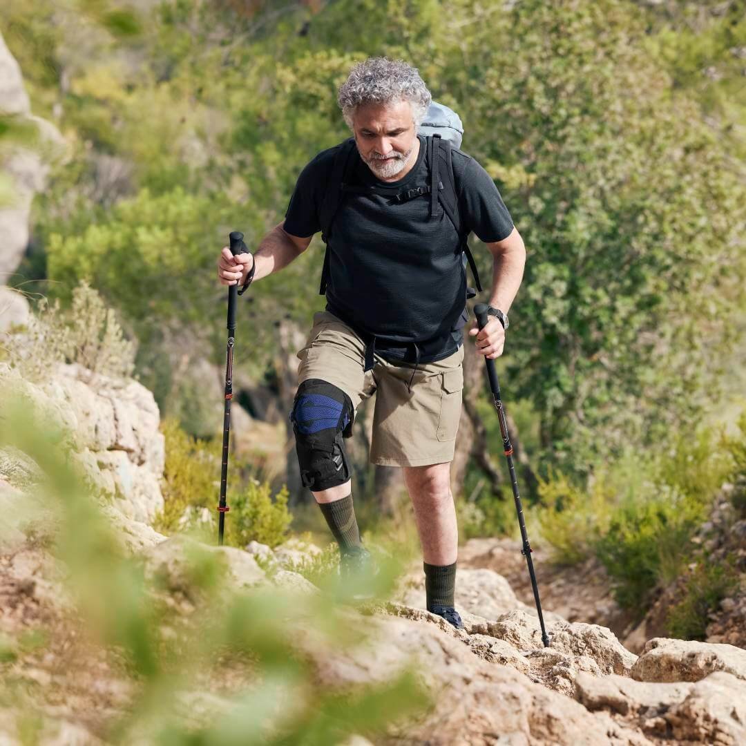 Älterer Mann mit Wanderausrüstung auf einem felsigen Pfad in einer grünen Landschaft, trägt die GenuTrain OA Knieorthese von Bauerfeind am rechten Bein zur Unterstützung beim Wandern.