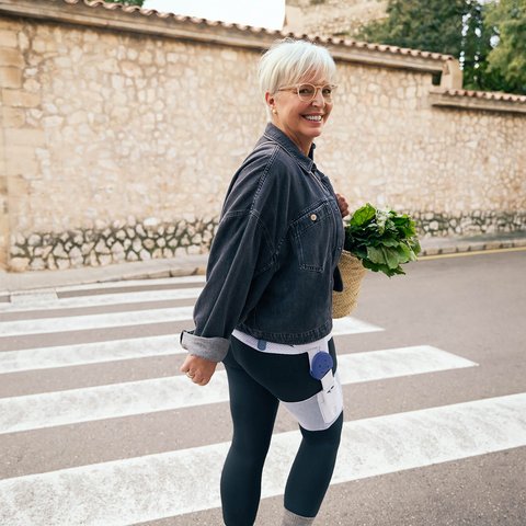 Eine Frau geht lächelnd über einen Zebrastreifen und blickt über die Schulter zur Kamera. Sie trägt eine schwarze Jeansjacke, eine Oberschenkelorthese am rechten Bein und hält eine Einkaufstasche mit frischem Gemüse in der linken Hand.