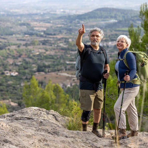 Ein älteres Paar steht mit Wanderstöcken auf einem Felsen und genießt den weiten Blick über eine grüne Hügellandschaft. Der Mann trägt eine GenuTrain OA Knieorthese zur Unterstützung bei Gonarthrose.