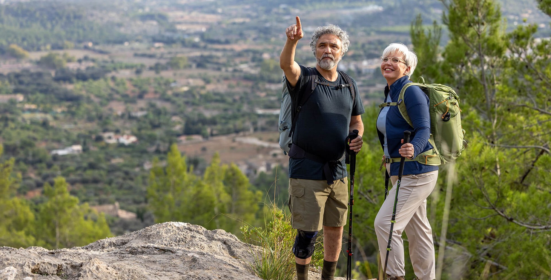 Ein älteres Paar steht mit Wanderstöcken auf einem Felsen und genießt den weiten Blick über eine grüne Hügellandschaft. Der Mann trägt eine GenuTrain OA Knieorthese zur Unterstützung bei Gonarthrose.