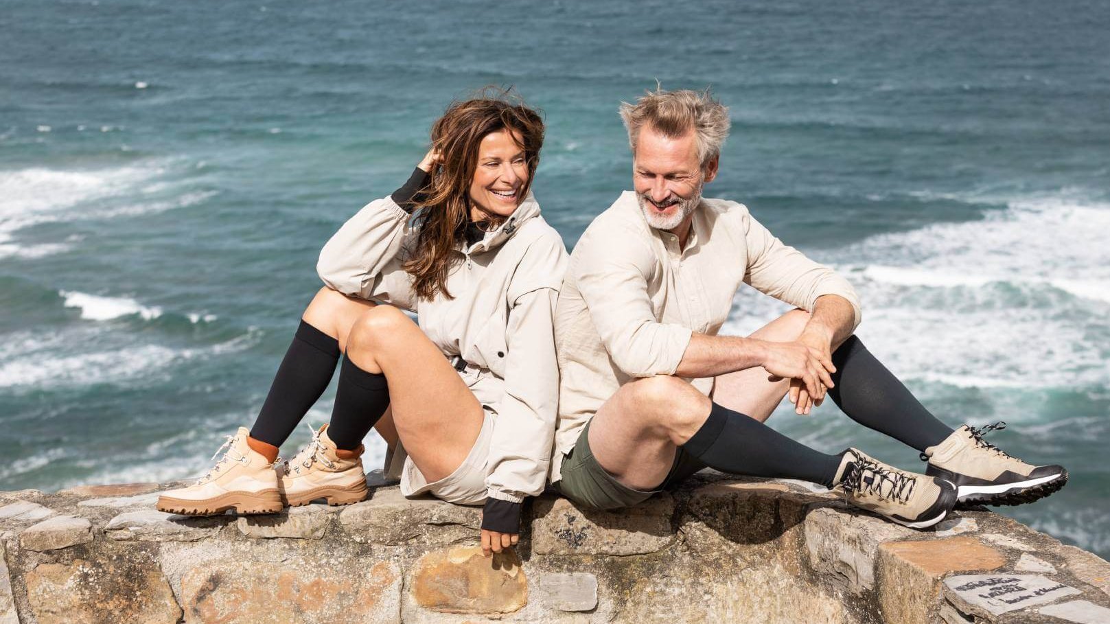 Ein Mann und eine Frau sitzen entspannt auf einer Steinmauer mit Blick auf das Meer im Hintergrund. Beide tragen Outdoor-Bekleidung und schwarze Kompressionsstrümpfe der Linie VenoTrain, kombiniert mit Wanderschuhen.