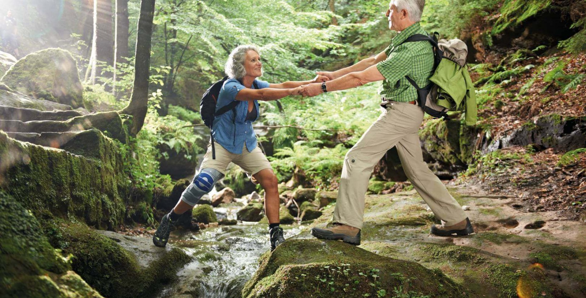 Das Bild zeigt ein Paar beim Wandern im Wald. Der Mann hilft seiner Frau über eine Fluss. Am Knie trägt die Wanderin eine SoftTec Genu von Bauerfeind zur Unterstützung.