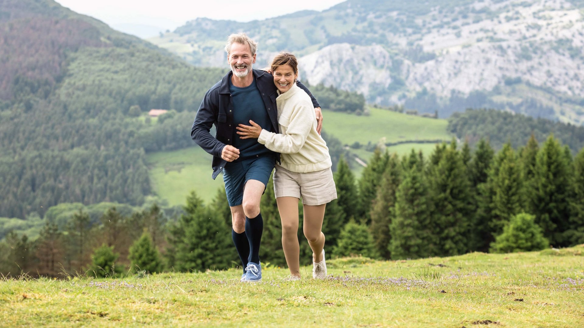 Ein Mann und eine Frau laufen Arm in Arm in einer Berglandschaft über eine Wiese. Beide tragen VenoTrain impuls Kompressionsstrümpfe von Bauerfeind. Diese helfen bei Besenreisern.