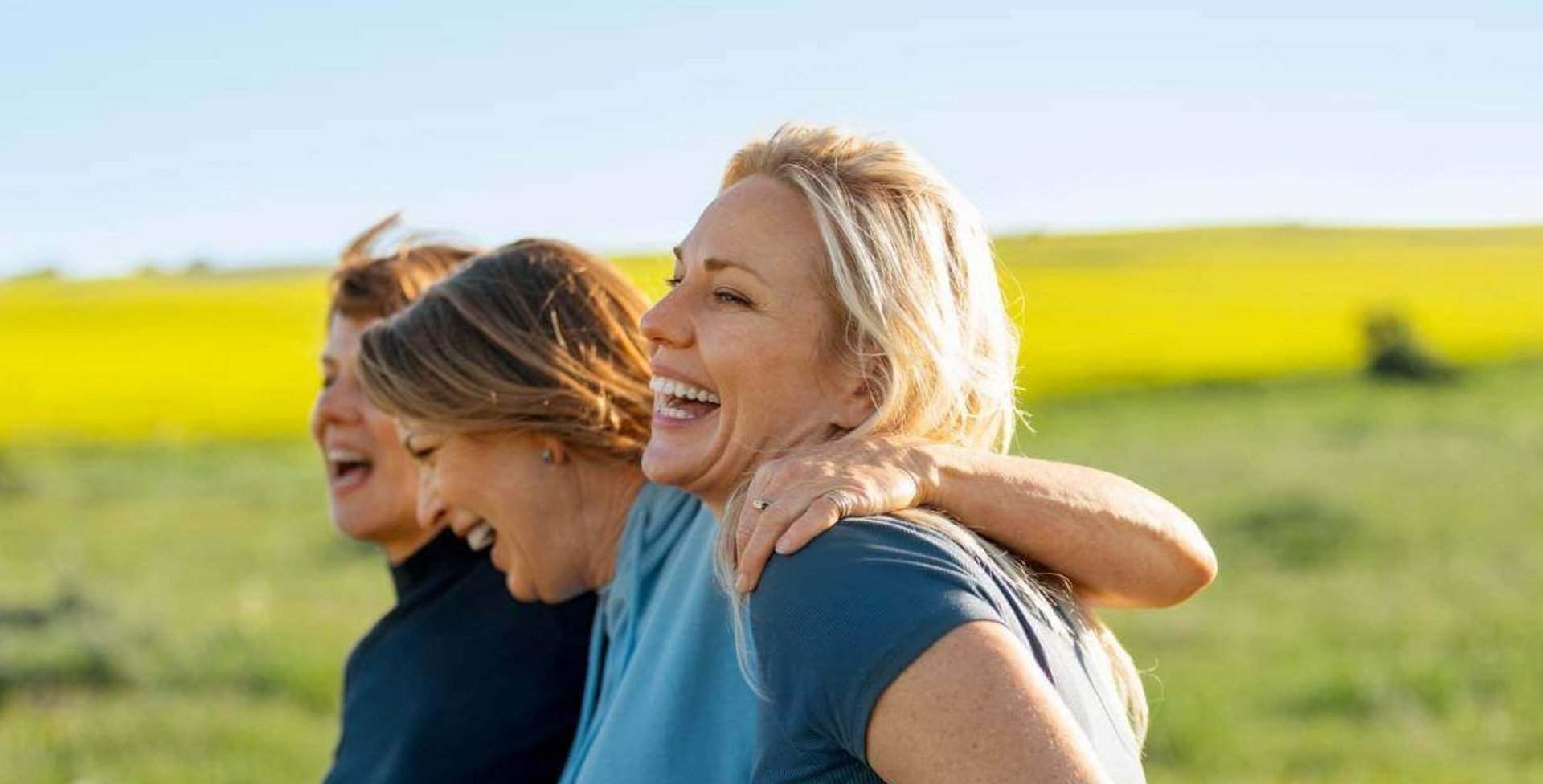 Das Bild zeigt, wie drei Frauen gut gelaunt und eng umschlungen auf einer frühlingshaften Wiese entlangspazieren.