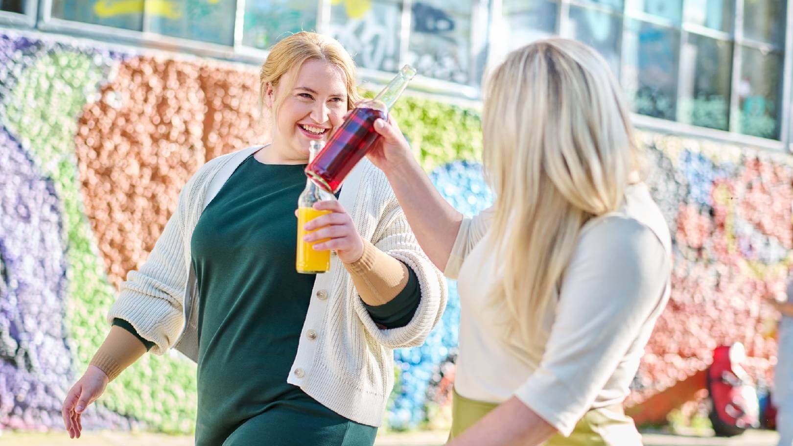 Zwei Frauen stoßen mit farbenfrohen Getränkeflaschen an, während sie lächelnd durch eine urbane Umgebung spazieren. Im Hintergrund ist eine bunte Wand mit grafischen Mustern zu sehen, die eine lebendige Atmosphäre schafft.