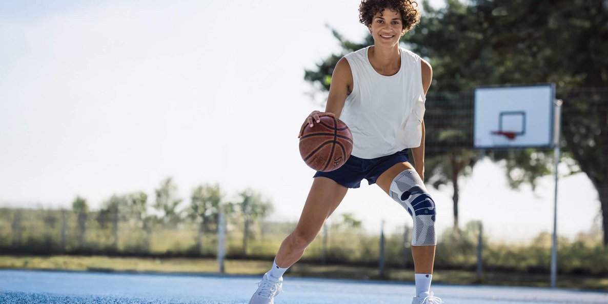 Junge Frau mit weissem Trägershirt, dunkelblauer Sporthose und weisser Kniebandage spielt Basketball auf einem Aussenplatz mit blauem Bodenbelag; sie dribbelt den Ball und lächelt in die Kamera, im Hintergrund ist ein Basketballkorb und Bäume sichtbar.