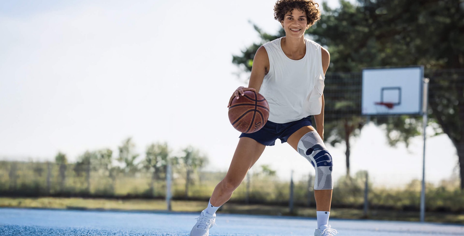 Junge Frau mit weissem Trägershirt, dunkelblauer Sporthose und weisser Kniebandage spielt Basketball auf einem Aussenplatz mit blauem Bodenbelag; sie dribbelt den Ball und lächelt in die Kamera, im Hintergrund ist ein Basketballkorb und Bäume sichtbar.