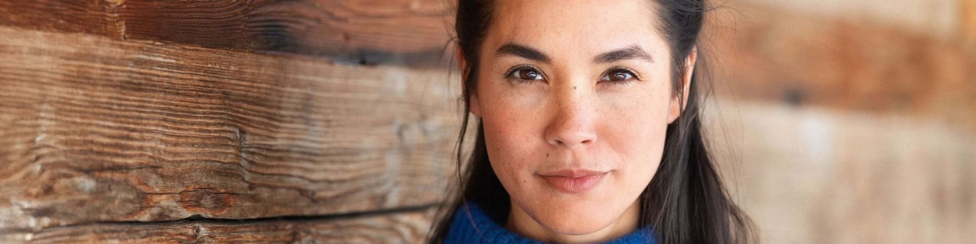 Eine junge Frau im blauen Pullover vor einer Holzwand, die in die Kamera blickt.