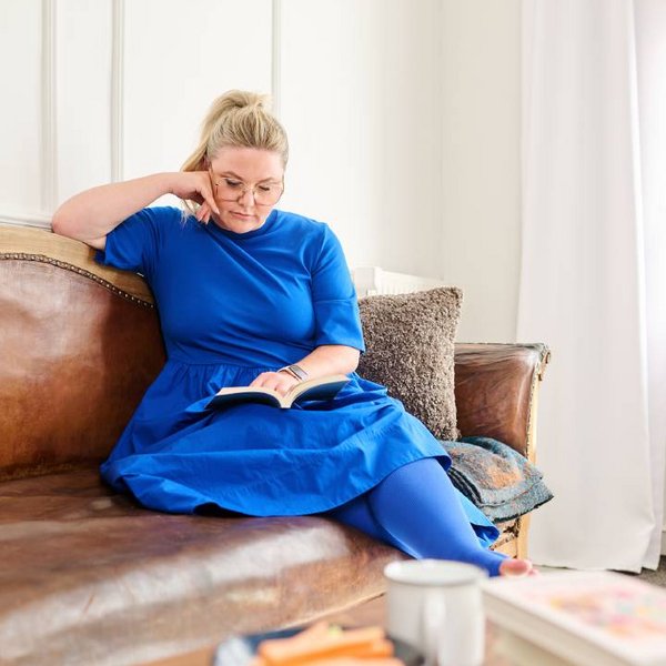 Eine Frau in einem leuchtend blauen Kleid sitzt auf einem Ledersofa und liest ein Buch. Sie trägt Kompressionsstrümpfe, die speziell für Lip- und Lymphödeme geeignet sind. Neben ihr auf dem Tisch stehen gesunde Snacks und eine Tasse. 