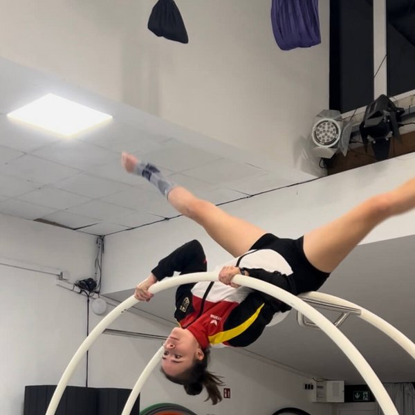 Larissa von Behren führt eine akrobatische Übung kopfüber im Rhönrad aus, trägt eine Sportjacke in Schwarz, Rot und Gelb sowie die AchilloTrain Achillessehnenbandage von Bauerfeind am rechten Fuß.