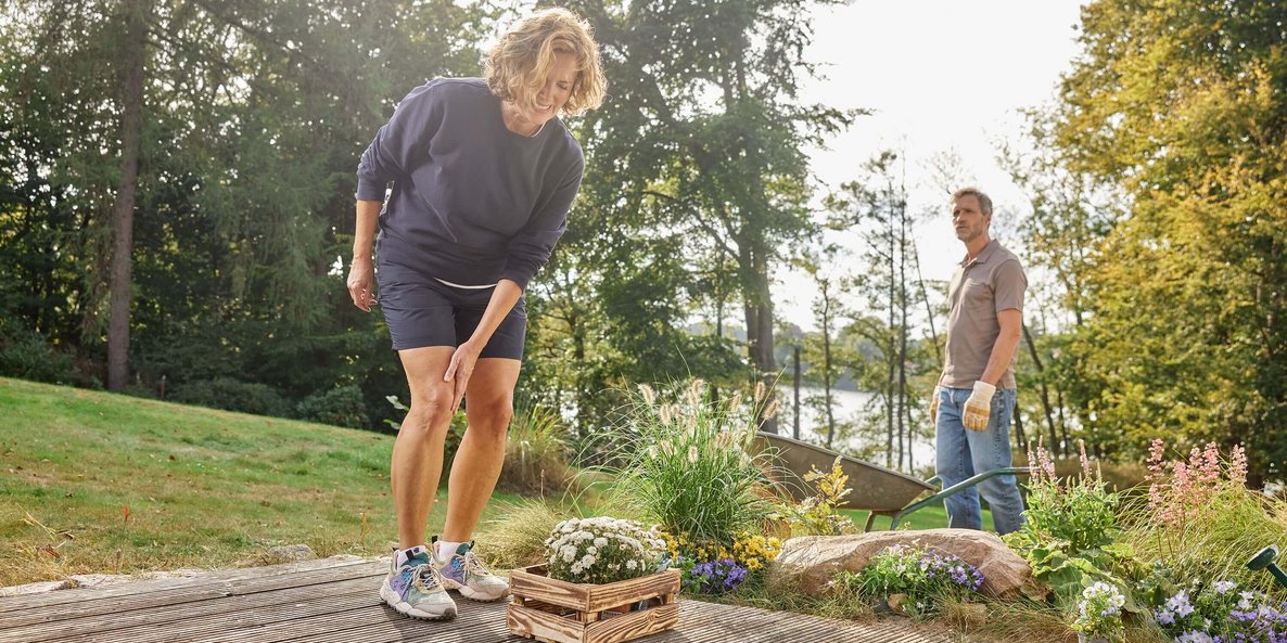 Eine Frau greift sich beim Gärtnern an ihr schmerzendes Knie. Im Hintergrund steht ein Mann mit Handschuhen und schaut zu. Der Garten ist von bunten Blumen und einer idyllischen Umgebung mit Bäumen und einem See geprägt.