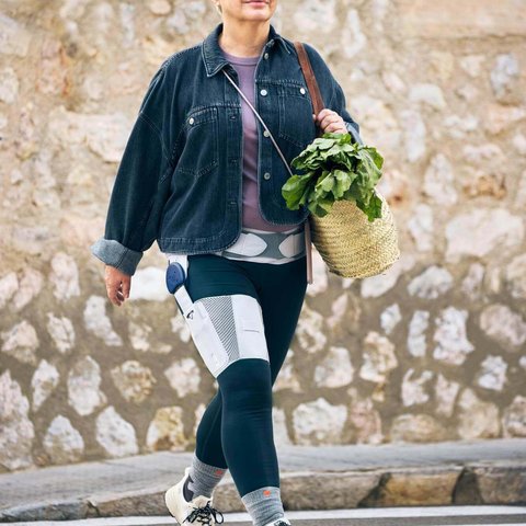 Eine ältere Frau mit kurzen weissen Haaren und Brille überquert einen Zebrastreifen. Sie trägt eine schwarze Jeansjacke, Leggings, weisse Turnschuhe und eine Orthese an der rechten Hüfte und am rechten Oberschenkel. In ihrer Hand hält sie eine geflochtene Tasche mit frischem Gemüse.