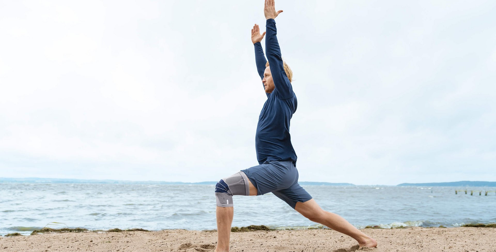 Das Bild zeigt einen Mann in Yoga-Pose am Strand. Am rechten Knie trägt er eine GenuTrain zur Unterstützung. Die Bandage bietet u.a. Unterstützung bei einem Kniegelenkserguss.