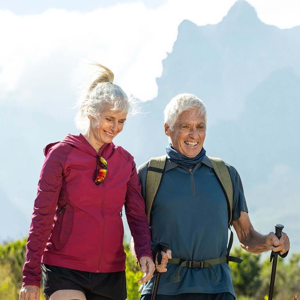  Auf dem Bild sieht man ein älteres Paar, das in einer malerischen Berglandschaft wandert. Die Frau trägt eine GenuTrain OA Orthese, die speziell für Menschen mit Arthrose entwickelt wurde, um ihr Kniegelenk zu unterstützen.