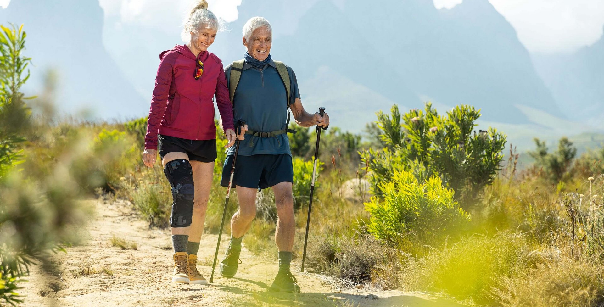  Auf dem Bild sieht man ein älteres Paar, das in einer malerischen Berglandschaft wandert. Die Frau trägt eine GenuTrain OA Orthese, die speziell für Menschen mit Arthrose entwickelt wurde, um ihr Kniegelenk zu unterstützen.