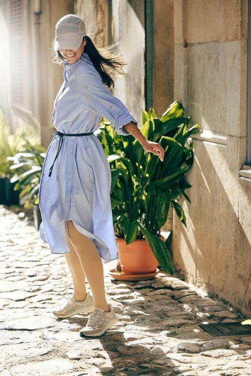 Eine lachende Frau mit wehenden Haaren dreht sich spielerisch in einer sonnigen Altstadtgasse. Sie trägt ein locker geschnittenes hellblaues Hemdblusenkleid, eine beigefarbene Cap und VenoTrain Kompressionsstrümpfe in einem natürlichen Ton, kombiniert mit sportlichen Sneakern.