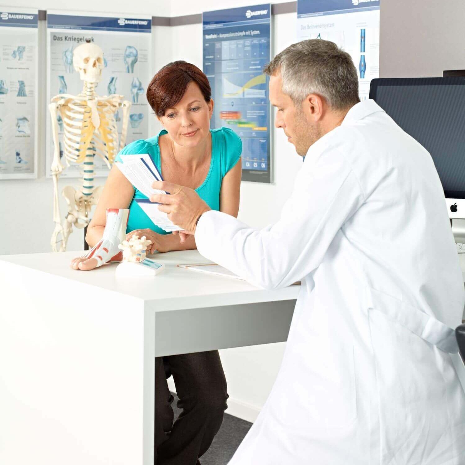 Das Bild zeigt eine Patientin zusammen mit einem Arzt an einem Tisch in einem Behandlungszimmer. Der Arzt spricht mit der Patientin über verschiedene Behandlungsmöglichkeiten des Sprunggelenks.