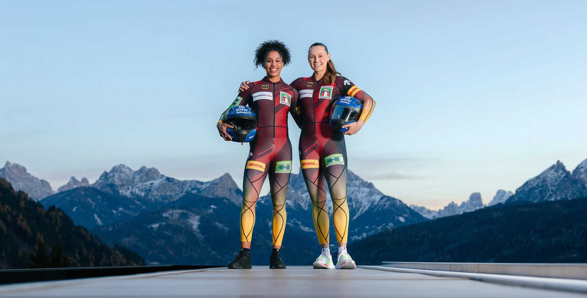 Zwei Bobfahrerinnen in eng anliegenden Rennanzügen mit den Farben und Logos der deutschen Nationalmannschaft stehen Arm in Arm auf einer Bobbahn mit verschneiten Alpen im Hintergrund. Beide halten ihre Helme in der Hand und lächeln in die Kamera.