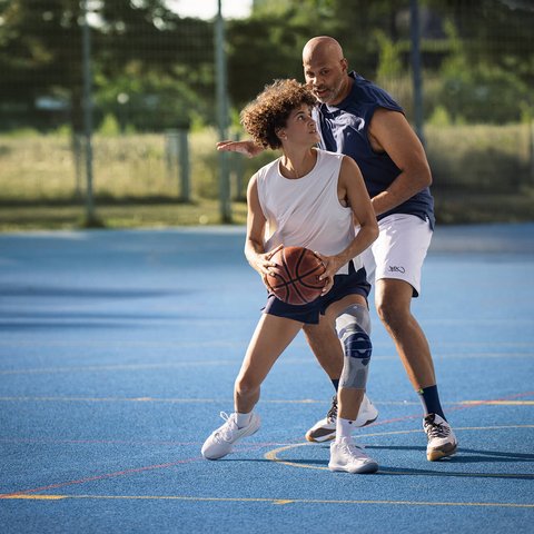 Zwei Personen spielen Basketball auf einem Aussenplatz mit blauem Boden. Die vordere Person mit weissem Trägershirt, dunklen Shorts und Kniebandage am rechten Bein hält den Ball in Spielposition, während die andere Person sie eng verteidigt.
