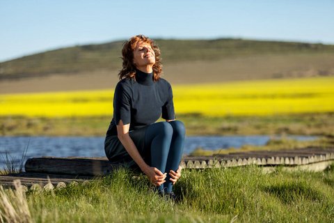 Eine Frau sitzt draußen auf einem Holzsteg, umgeben von einer natürlichen Landschaft mit gelben Blumenfeldern und einem See im Hintergrund. Sie trägt eine dunkelblaue Kompressionsstrumpfhose und ein passendes Oberteil, ihre Haare wehen leicht im Wind. 
