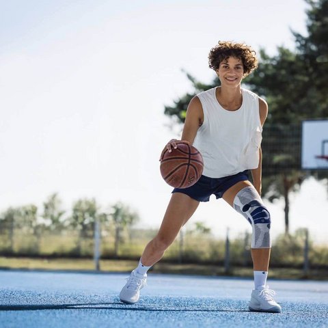 Eine Person mit weissem Trägershirt, dunklen Shorts und Kniebandage am rechten Bein dribbelt einen Basketball auf einem blauen Aussenplatz. Im Hintergrund ist ein Basketballkorb und ein Zaun sichtbar. Die Person lächelt und schaut in die Kamera.