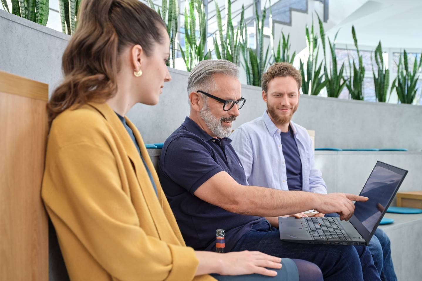 Drei Personen sitzen in einer modernen, begrünten Arbeitsumgebung. Ein Mann zeigt auf den Bildschirm eines Laptops, während die anderen aufmerksam zuhören. Die Szene vermittelt eine entspannte und produktive Atmosphäre.