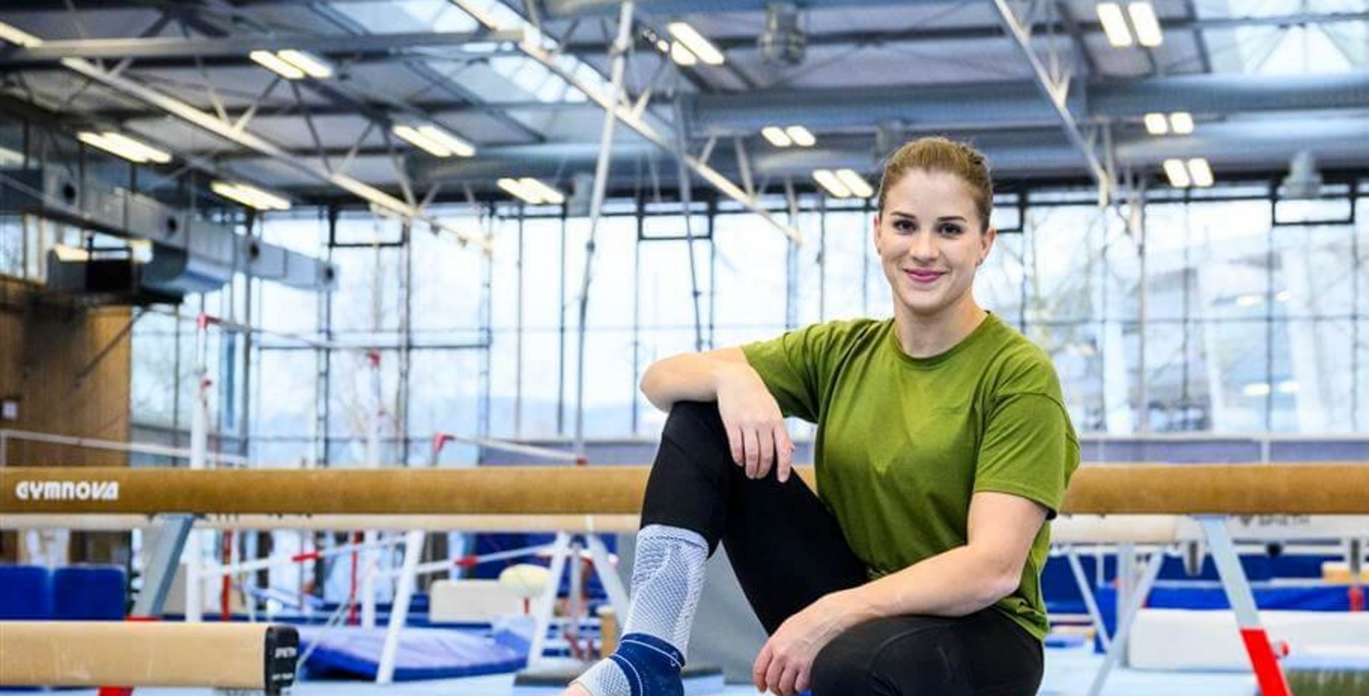 Turnerin Elisabeth Seitz sitzt auf einem Holzkasten in einer Sporthalle, trägt ein grünes T-Shirt, schwarze Leggings und die AchilloTrain-Bandage von Bauerfeind am rechten Fuß, umgeben von Turngeräten wie Schwebebalken.