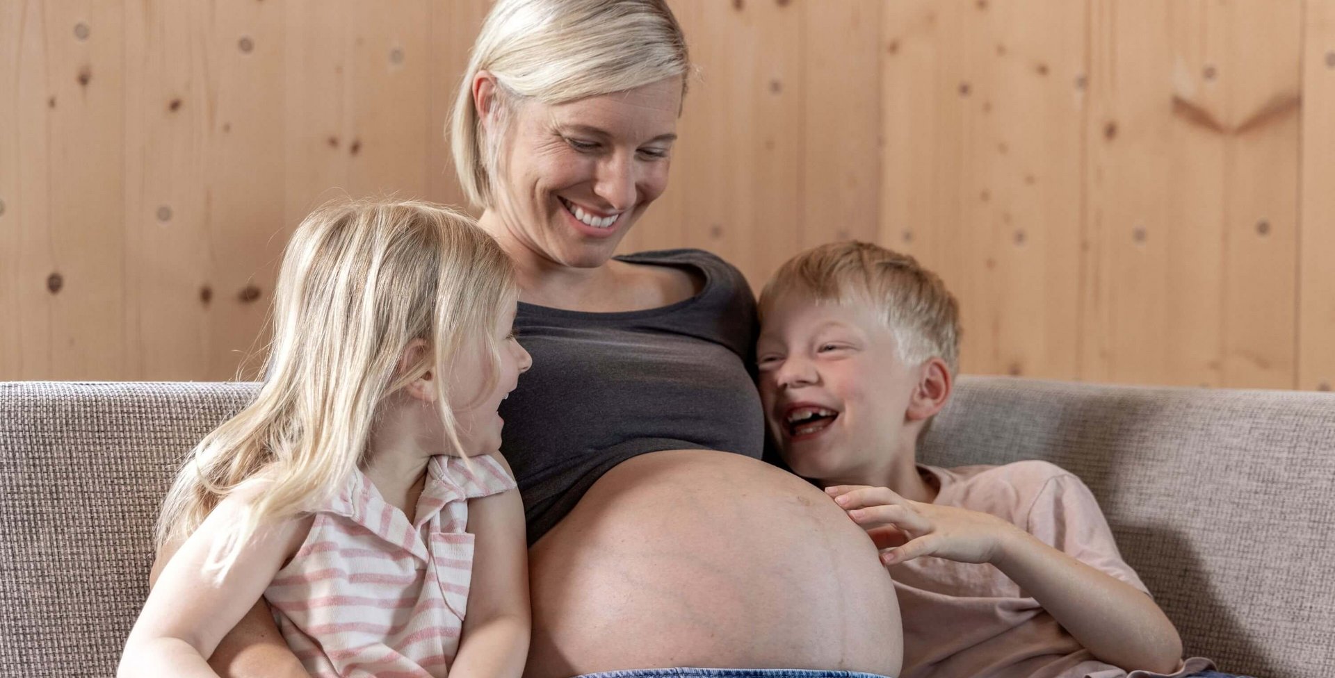  Schwangere Frau sitzt auf einem Sofa und lächelt, während ihre beiden Kinder fröhlich ihren Babybauch berühren.