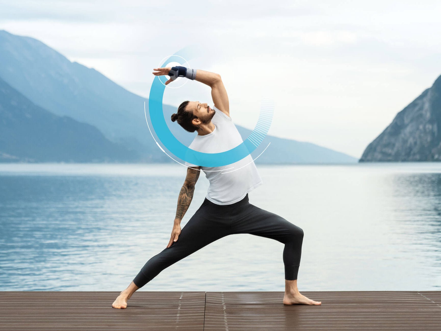 Person in Sportkleidung trägt Bauerfeind Hand-Bandage ManuTrain und macht Yoga-Pose auf Holzsteg vor Bergsee mit bewölktem Himmel.