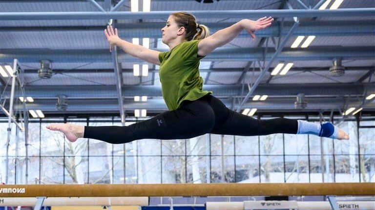 Turnerin Elisabeth Seitz führt in einer Sporthalle einen eleganten Sprung über den Schwebebalken aus, trägt ein grünes T-Shirt, schwarze Leggings und eine AchilloTrain Bandage am rechten Fuß.
