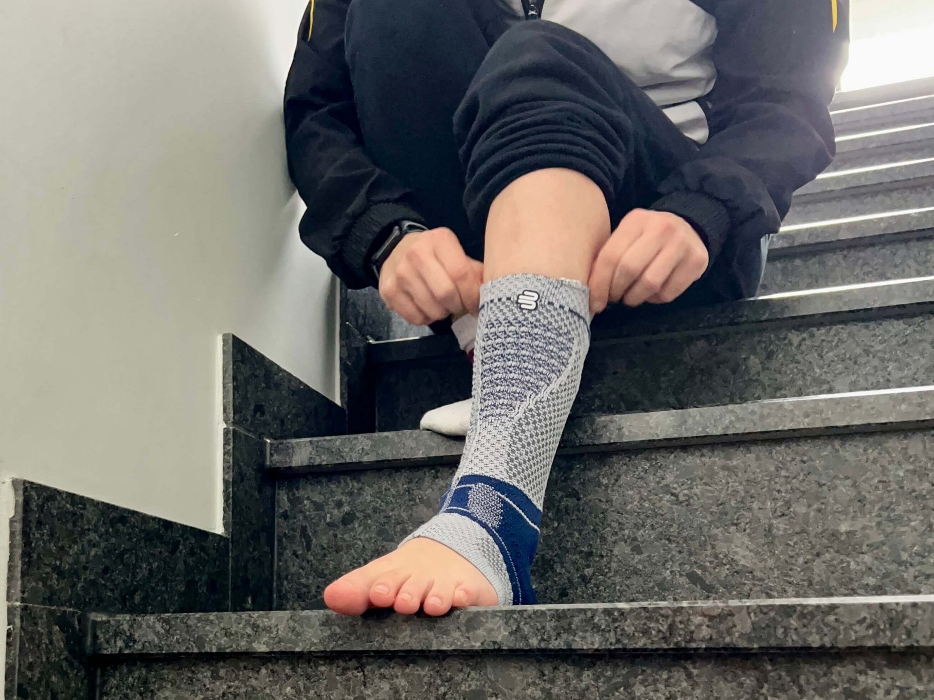 Rhönrad-Sportlerin Larissa von Behren sitzt auf einer Treppe und zieht die AchilloTrain Achillessehnenbandage von Bauerfeind am rechten Fuß an, bekleidet mit einer Sportjacke und Trainingshose.