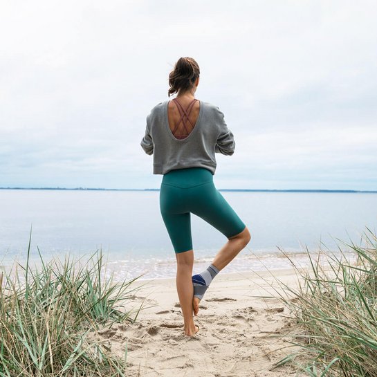 Das Bild zeigt eine Frau, die am Strand eine Yoga-Pose einnimmt. Sie trägt die MalleoTrain Sprunggelenkbandage von Bauerfeind, um ihr Sprunggelenk zu stabilisieren. Diese kommt u.a. bei einer Sprunggelenksdistorsion zum Einsatz.