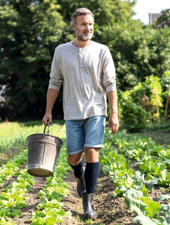 Ein Mann trägt VenoTrain hero Kompressionsstrümpfe in Dunkelblau und arbeitet im Garten. Er hält einen Metalleimer und trägt Gummistiefel sowie eine lässige Kleidungskombination aus grauem Langarmshirt und Jeansshorts. Im Hintergrund sind grüne Pflanzen und Bäume zu sehen.