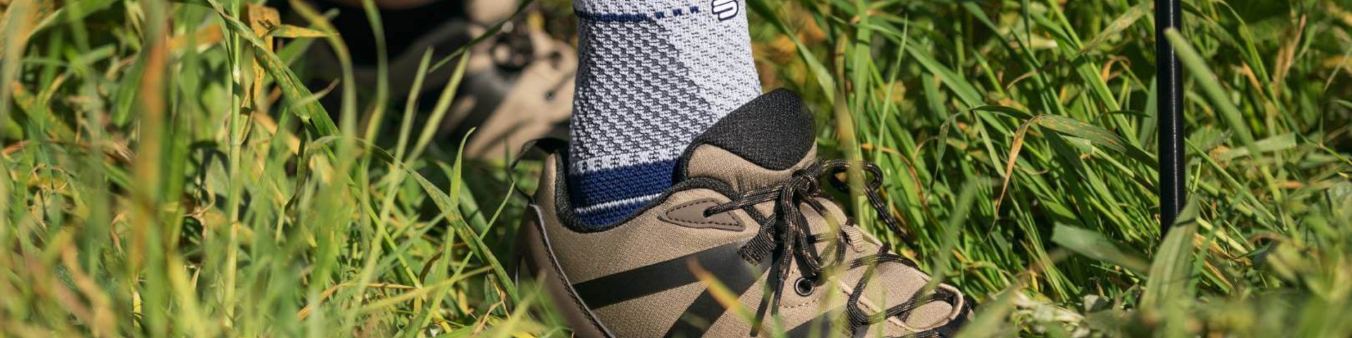 Nahaufnahme eines Fußes in einem Wanderschuh, der eine MalleoTrain Sprunggelenkbandage von Bauerfeind trägt, stehend im hohen Gras mit einem Wanderstock im Hintergrund.