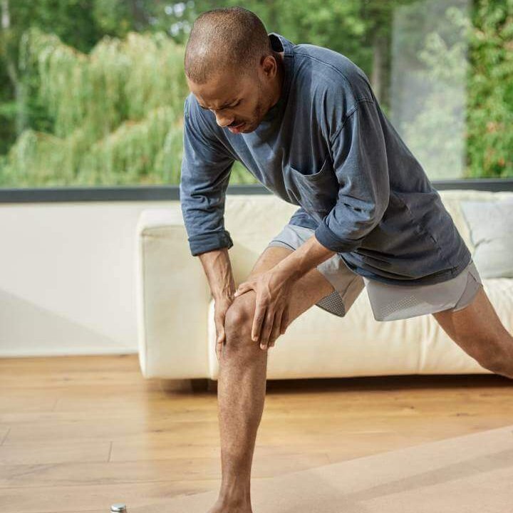 Ein Mann greift sich während des Trainings an das schmerzende Knie. Er trägt sportliche Kleidung und befindet sich in einem modernen Raum mit großen Fenstern. Im Vordergrund sind eine Wasserflasche, ein Springseil und eine Yoga-Matte zu sehen.