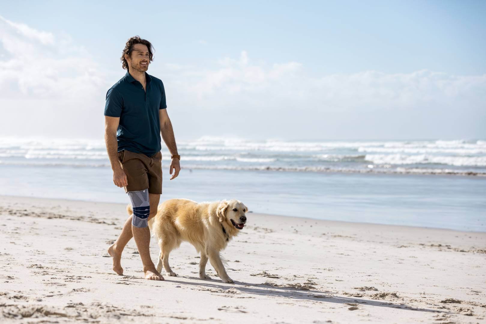 banner-knieschmerzen Ein Mann spaziert barfuß mit einem Golden Retriever an einem sonnigen Sandstrand. Er trägt eine GenuTrain Kniebandage von Bauerfeind, während im Hintergrund Wellen das Ufer erreichen.