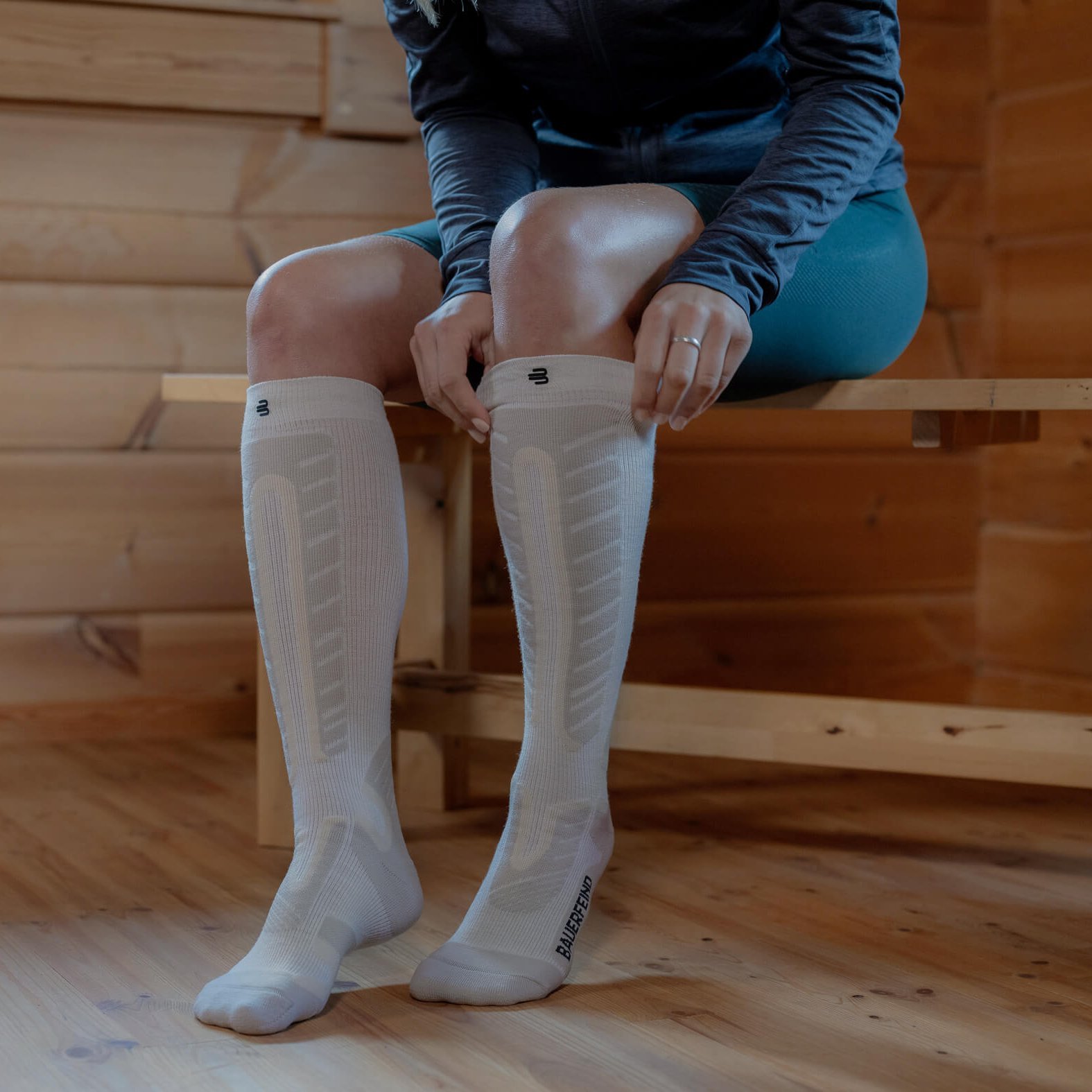 Person zieht weiße Sports Alpine Compression Socks von Bauerfeind an, während sie auf einer Holzbank in einem Raum mit Holzvertäfelung sitzt. Die Strümpfe bieten eine gezielte Kompression für sportliche Aktivitäten.