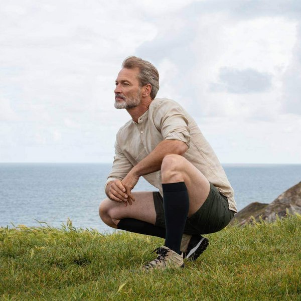 Ein Mann hockt auf einer Wiese am Meer. Er trägt Kompressionsstrümpfe von Bauerfeind.