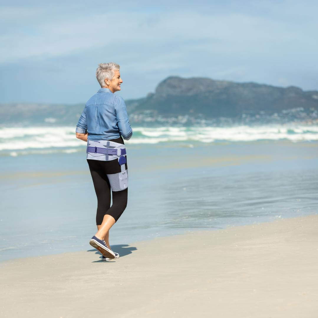  Abgebildet ist eine ältere Frau, die am Strand entlang spaziert. Sie trägt eine CoxaTrain Orthese, die speziell für Patienten mit Hüftarthrose entwickelt wurde.