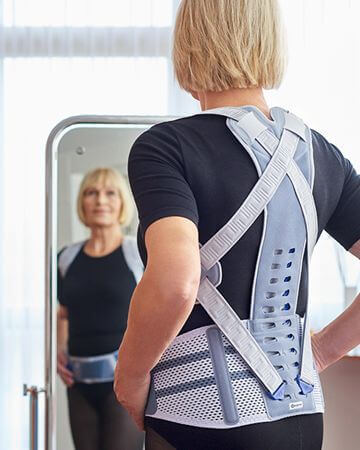 Eine Frau mit Rückenorthese von Bauerfeind, der Spinova Osteo, betrachtet sich im Spiegel.