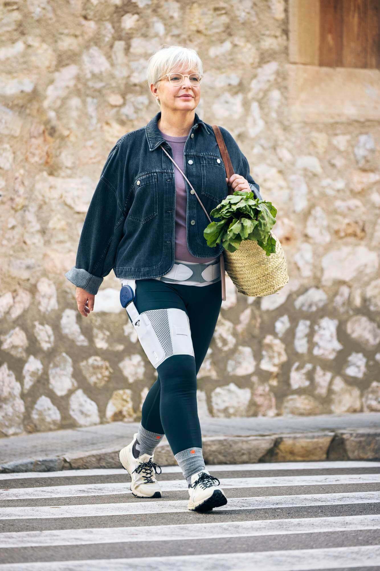 Eine ältere Frau mit kurzen weissen Haaren und Brille überquert einen Zebrastreifen. Sie trägt eine schwarze Jeansjacke, Leggings, weisse Turnschuhe und eine Orthese an der rechten Hüfte und am rechten Oberschenkel. In ihrer Hand hält sie eine geflochtene Tasche mit frischem Gemüse.