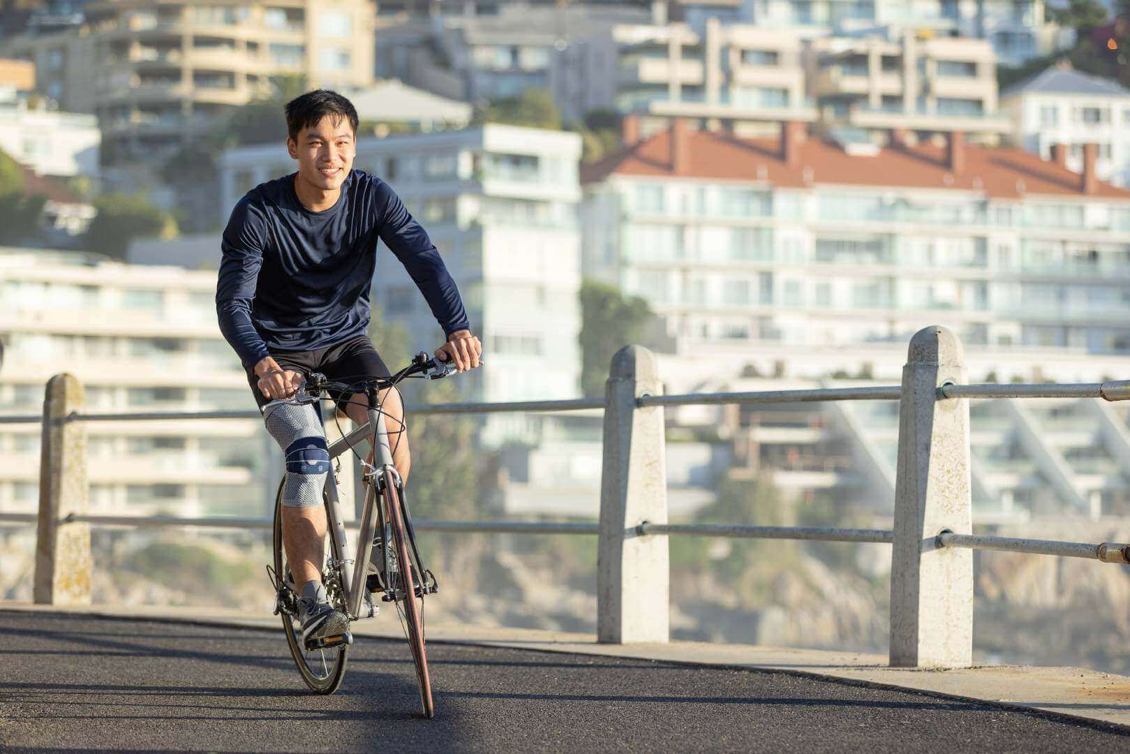 Das Bild zeigt einen Mann, der mit einem Fahrrad an einer Strandpromenade entlangfährt und dabei eine GenuTrain Bandage trägt, um seine Kniebeschwerden zu lindern. Radfahren erweist sich als gelenkschonende Sportart.