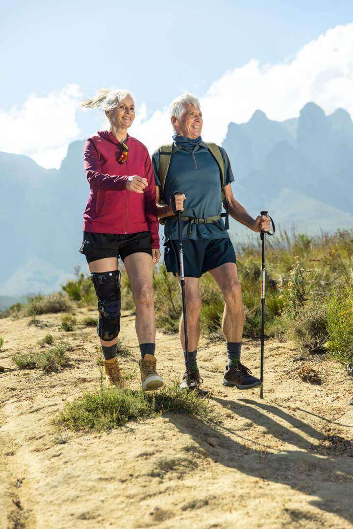Abgebildet ist ein Seniorenpärchen beim Wandern in einer sommerlichen Berglandschaft. Zur Unterstützung ihres Kniegelenks trägt die Frau eine GenuTrain OA Orthese, die speziell für Arthrose-Patienten entwickelt wurde.