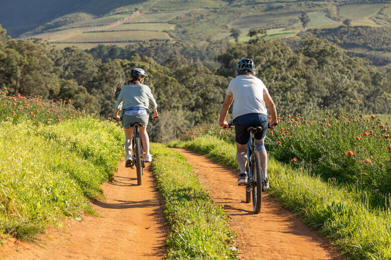 Zwei Personen fahren auf einem roten Sandweg mit Mountainbikes durch eine malerische Landschaft. Beide tragen Fahrradhelme und sportliche Kleidung. Der Weg ist von grüner Vegetation und Blumen gesäumt. 
