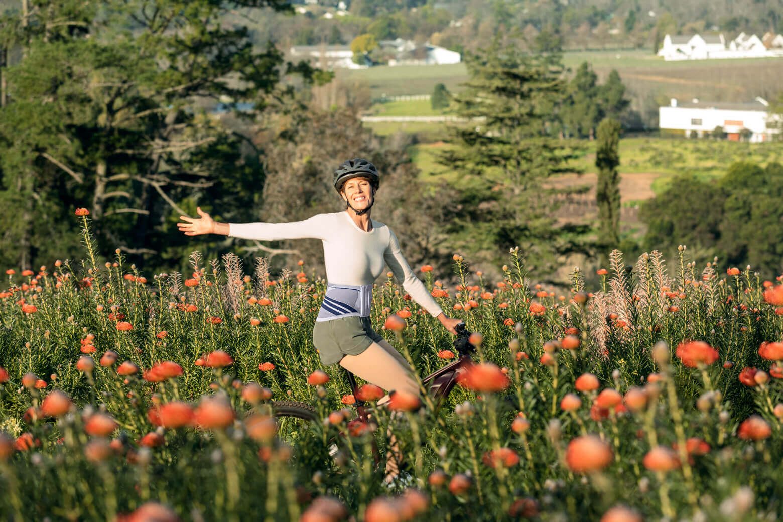  Eine Frau steht mit ausgebreiteten Armen auf einer Wiese voller orangefarbener Blumen. Sie trägt einen Fahrradhelm, eine LumboTrain Rückenbandage sowie sportliche Kleidung und hält ein Fahrrad. 