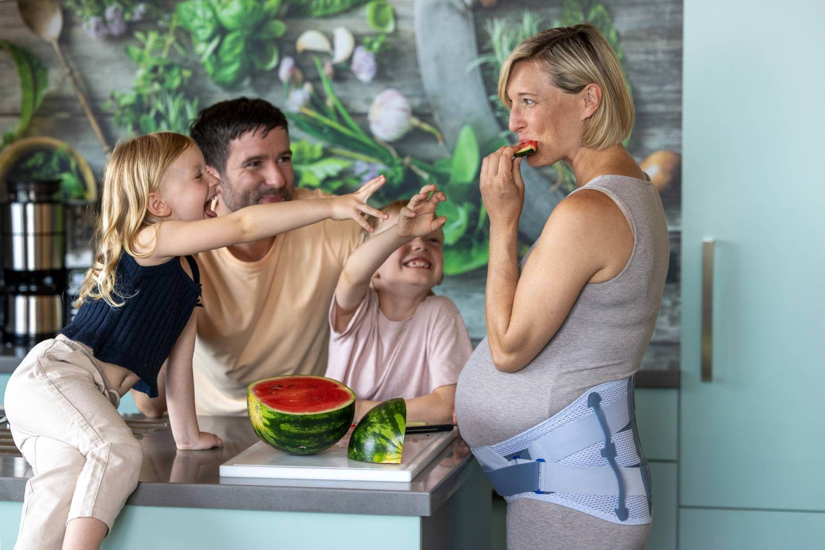 Eine schwangere Frau mit Spinova Mum Orthese isst ein Stück Wassermelone in der Küche, während ihre Tochter lachend nach einem Stück greift. Der Vater und der Sohn schauen fröhlich zu.