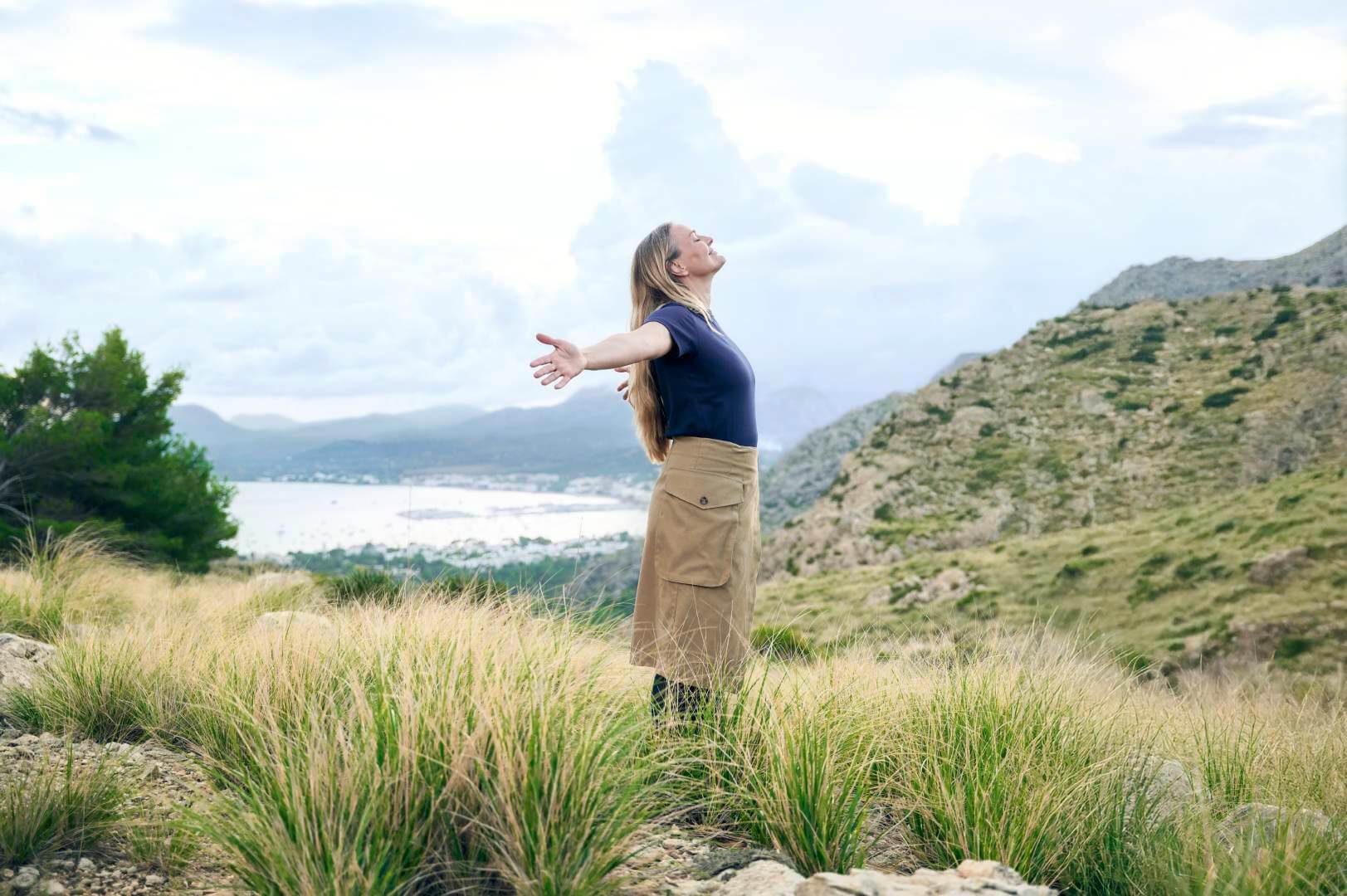 Eine Frau mit langen, offenen Haaren steht auf einem Hügel mit Blick auf eine malerische Küstenlandschaft. Sie breitet die Arme aus, atmet tief ein und genießt die frische Bergluft. Ihr Outfit in Erdtönen ergänzt den natürlichen Look, während dezente VenoTrain Kompressionsstrümpfe für Komfort sorgen.